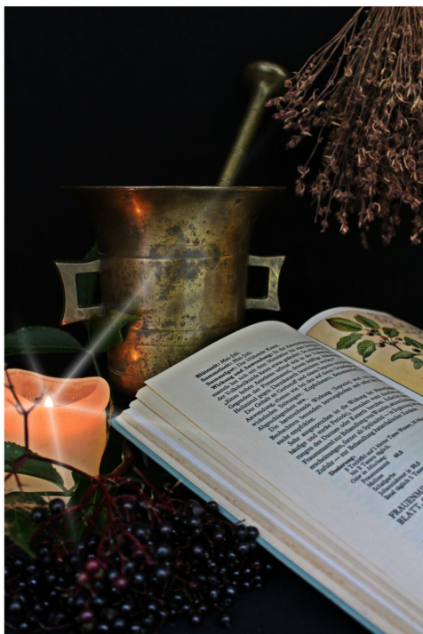 Dark purple berries, candle, copper pot, dried herbs, and open book against black background