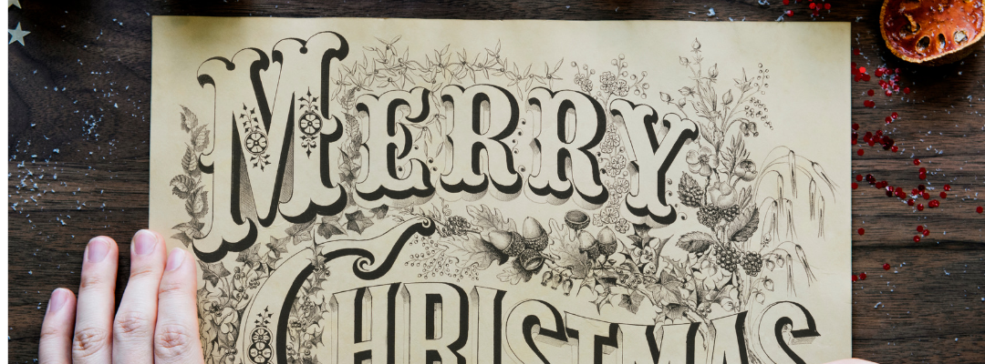 Pair of hands holding a pen over an ornate sign reading "Merry Christmas, surrounded by pine boughs & frosted pine cones